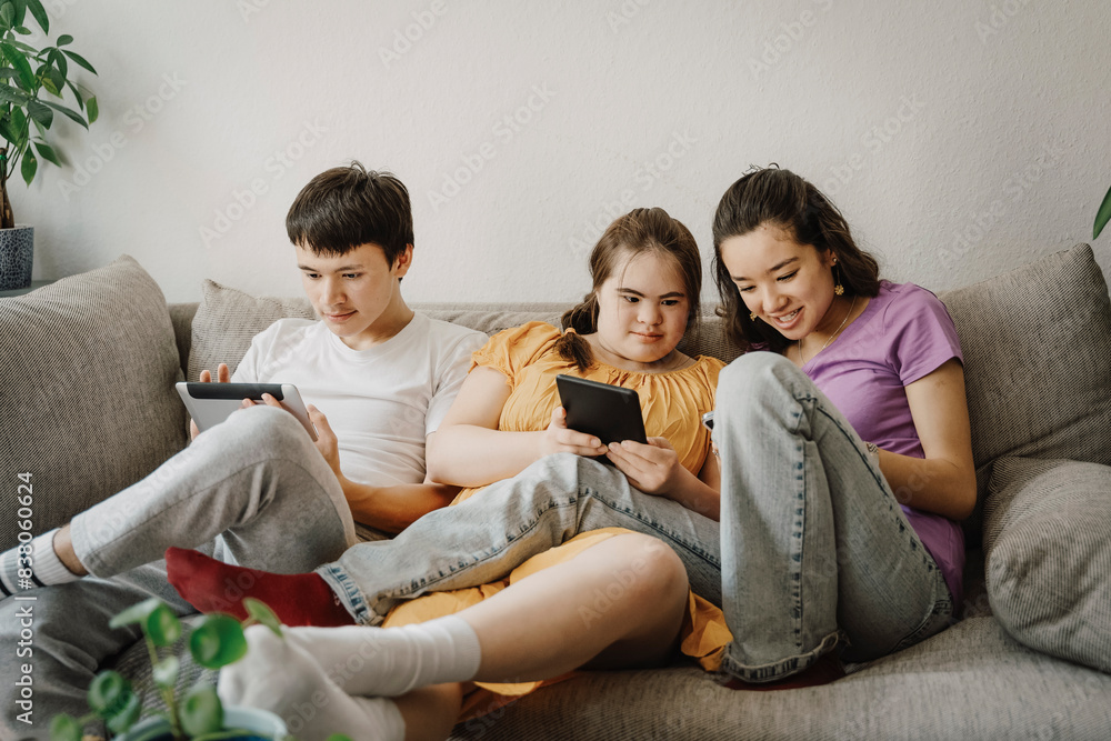 Curious siblings using wireless technologies with down syndrome sister sitting on sofa at home Curious siblings using wireless technologies with down syndrome sister sitting on sofa at home