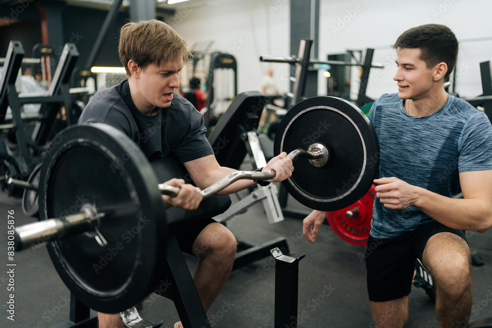 Exhausted male exercising support under supervision of personal coach. Trainer helping beginner sportsman for correct doing Scott Curls with barbell, lifting up EZ-bar sitting on preacher curl bench. Exhausted male exercising support under supervision of personal coach. Trainer helping beginner sportsman for correct doing Scott Curls with barbell, lifting up EZ-bar sitting on preacher curl bench.