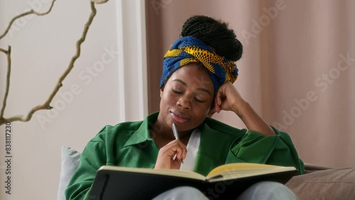 African American girl sits on couch alone, holding notebook and writing thoughts down in diary. Female black student taking notes or young writer working on a new story. She ponders and thinks over.