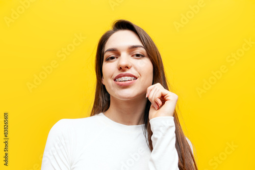 young woman with braces smiling on yellow isolated background, cheerful girl holding hand on hair and looking at camera
