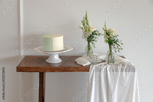 A beautifully decorated cake on a white stand sits on a rustic wooden table adorned with elegant vases of white flowers and flowing fabric, creating a serene and minimalist celebration setup.