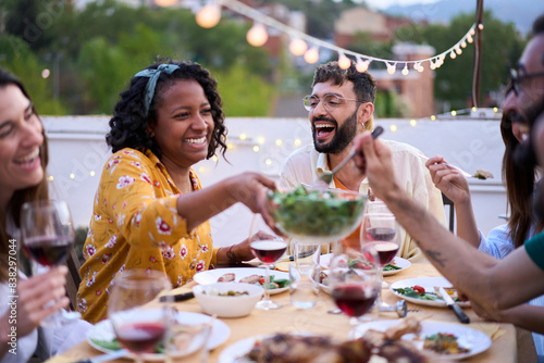 Smiling African American woman sitting at table passing salad to happy friends at meal event outdoors. Multiracial cheerful young people enjoying BBQ reunion together at weekend on rooftop at dusk