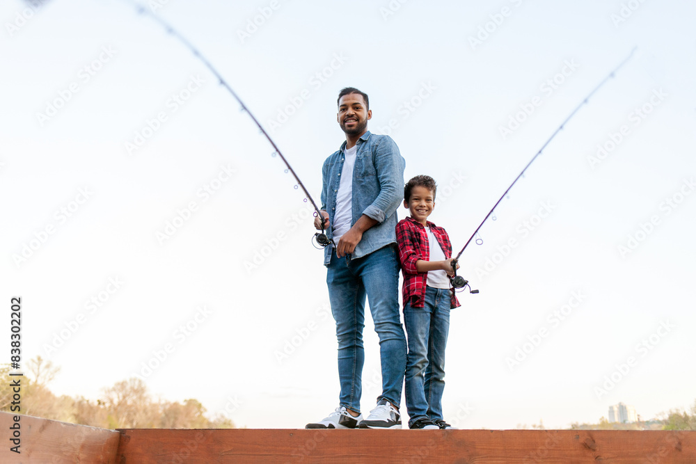 African American man and child standing on wooden pier holding fishing rods on the river, dad teaching his son to fish in the lake, family resting and relaxing on the weekend African American man and child standing on wooden pier holding fishing rods on the river, dad teaching his son to fish in the lake, family resting and relaxing on the weekend