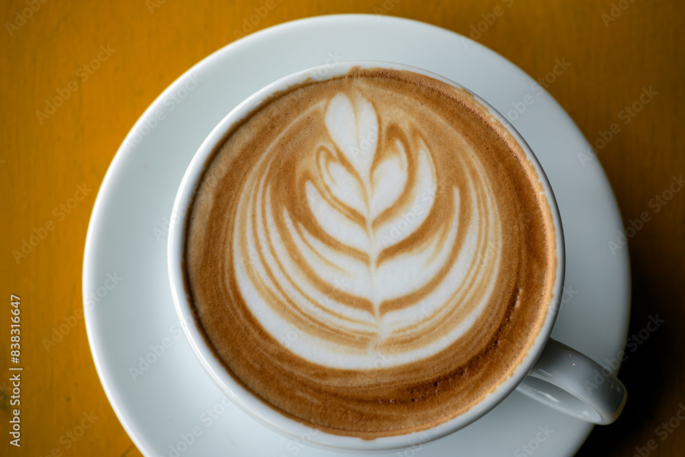 A detailed close-up of latte with intricate leaf art in white cup, set on yellow wooden table A detailed close-up of latte with intricate leaf art in white cup, set on yellow wooden table