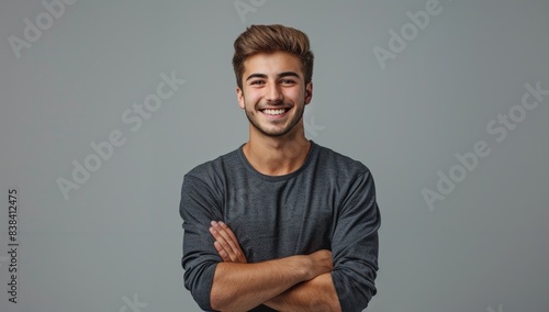 A handsome young man smiling with his arms crossed against a grey background, wearing casual with a confident and happy expression in a studio portrait.