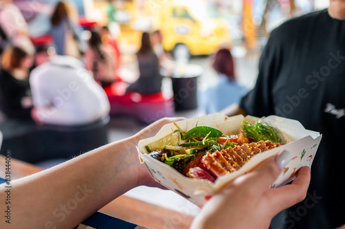 Hands holding a paper container filled with Asian street food  featuring noodles and vegetables  against a blurred background of a busy market