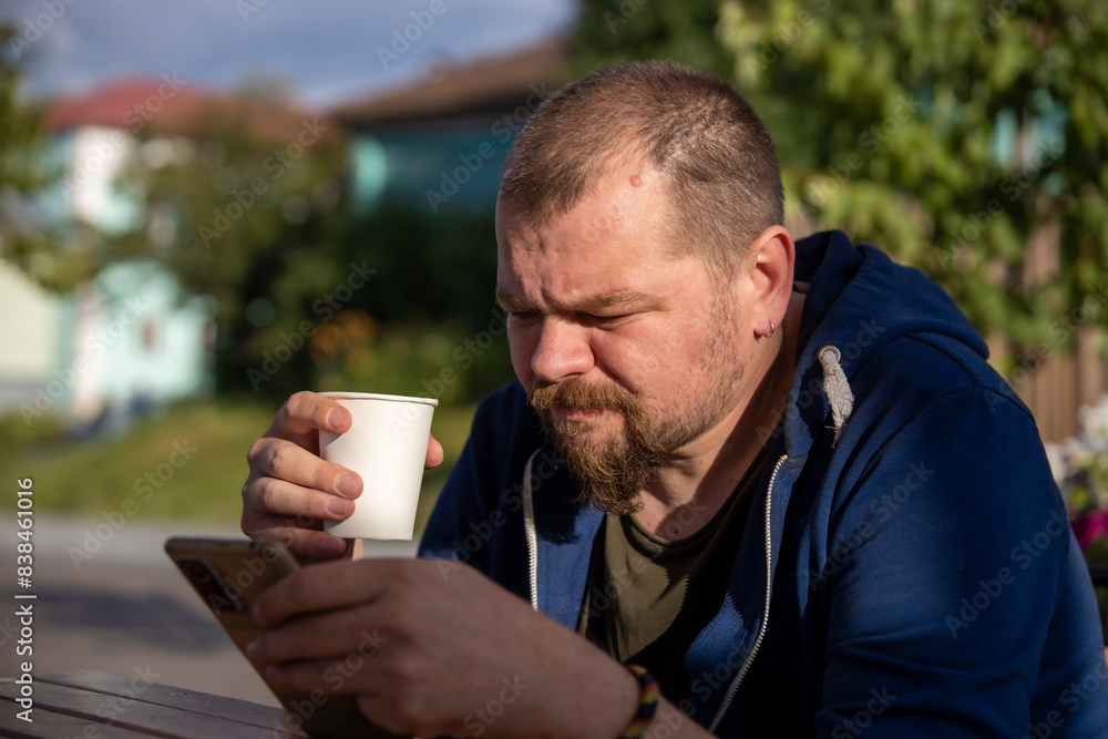 A bearded man sits at a table outside, enjoying a cup of coffee and scrolling through his phone. The sun is shining and the surrounding greenery suggests a warm summer day. A bearded man sits at a table outside, enjoying a cup of coffee and scrolling through his phone. The sun is shining and the surrounding greenery suggests a warm summer day.