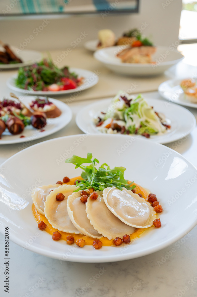 A white plate with round ravioli. The plate is set on a table with other plates of food in the background. A white plate with round ravioli. The plate is set on a table with other plates of food in the background.
