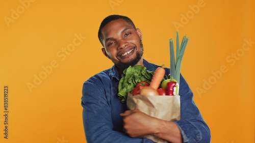Male model hugging his paper bag full of organic homegrown food, enjoying heathy eating from local bio supermarket. Young vegan person recommending ethically sourced produce. Camera A. photo