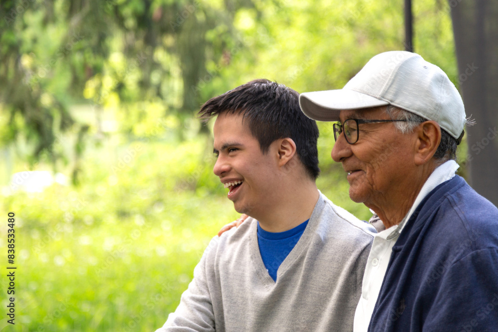A man and a young man are sitting in a park. The young man is wearing a blue shirt and a gray sweater. Both are smiling and laughing. A man and a young man are sitting in a park. The young man is wearing a blue shirt and a gray sweater. Both are smiling and laughing.