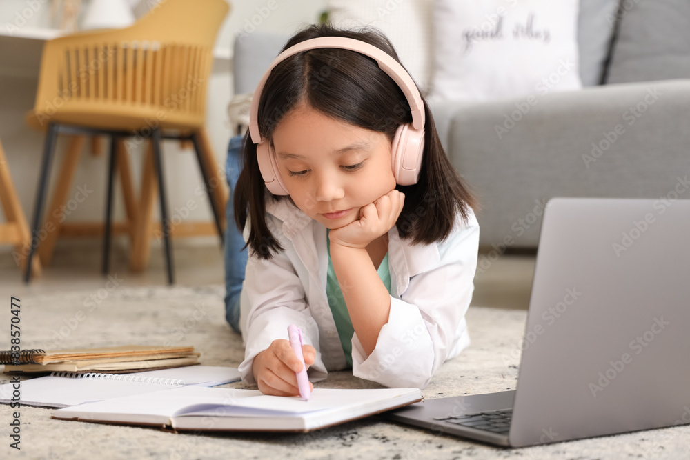 Little Asian girl in headphones with notebook and laptop studying online at home