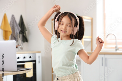 Little Asian girl with headphones and laptop dancing in kitchen