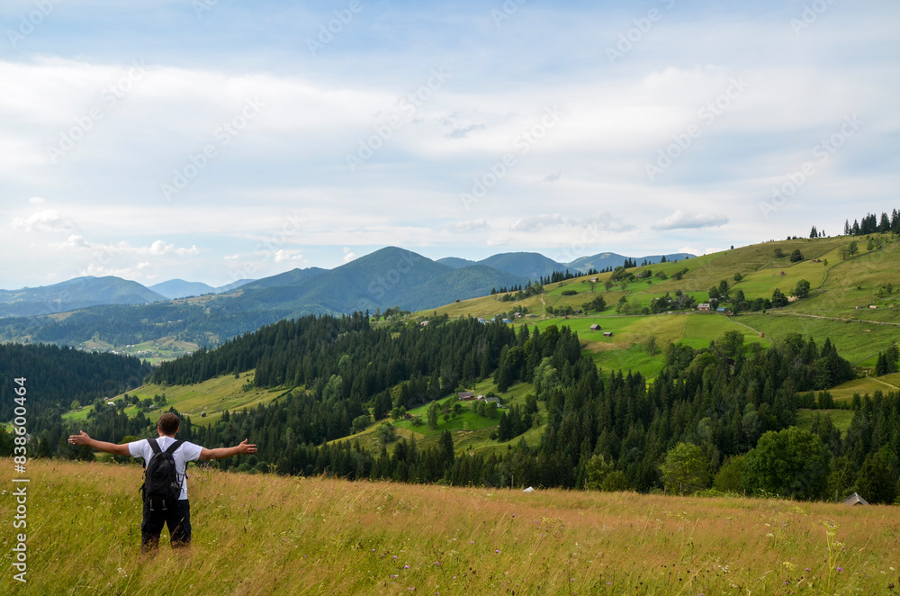 Man hiker with backpack stands with outstretched arms amidst a vast, serene landscape of rolling green hills, lush forests, and distant mountains under a partly cloudy sky. Carpathians, Ukraine