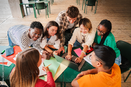 Big group of multiracial teenage students working together on the homework at the high school library. Teamwork of diverse classmates learning doing the class task with a laptop at university campus.