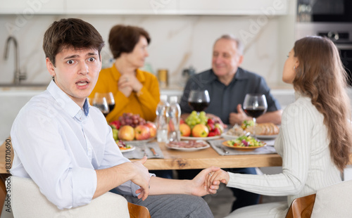 Shocked concerned young man sitting at table during family conversation with elderly parents at dinner  while girlfriend providing comfort by holding hand