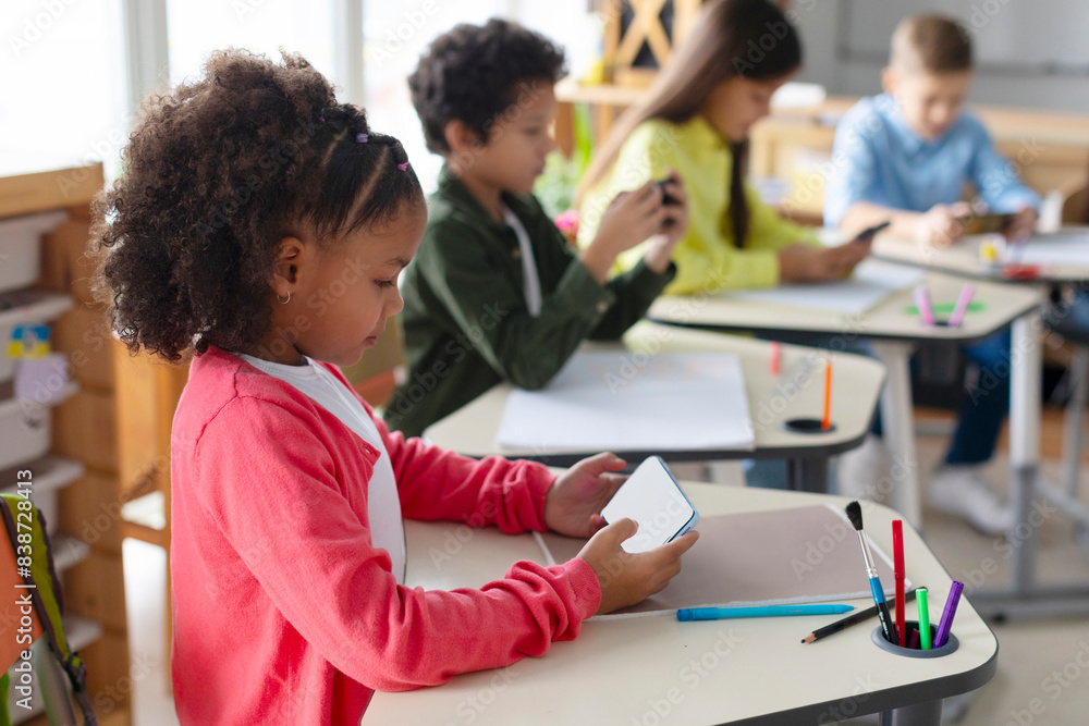 Diverse pupils schoolkids using smartphones in classroom, having break after lessons, children holding devices, playing games Diverse pupils schoolkids using smartphones in classroom, having break after lessons, children holding devices, playing games