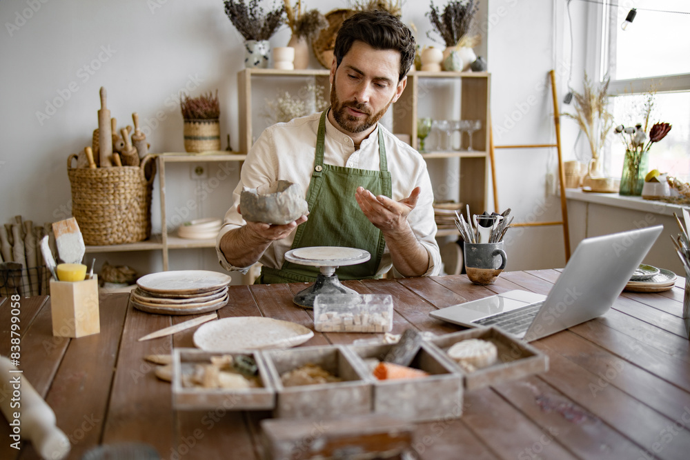 Handsome mature male potter in apron holding online lesson on clay vase sculpting using laptop. Bearded male studying remotely at university sitting in pottery studio. Handsome mature male potter in apron holding online lesson on clay vase sculpting using laptop. Bearded male studying remotely at university sitting in pottery studio.