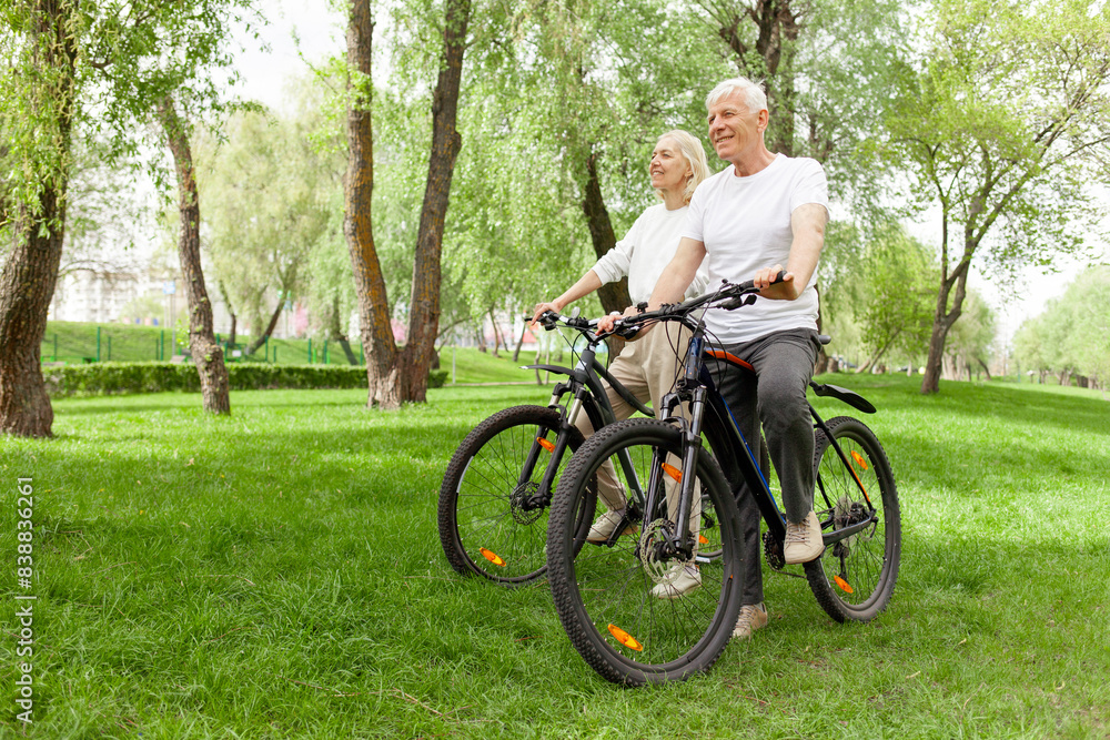 Obraz premium elderly senior couple rides bicycle in the park in the summer and smiles, old gray-haired man and woman are actively resting outdoors, old people practice cycling in forest
