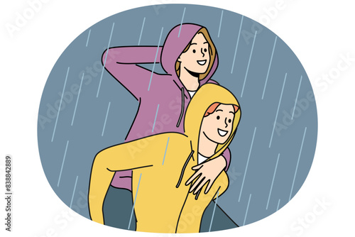 Happy couple walks in rain enjoying downpour after long drought and not wanting to go home. Man in waterproof hood rolls laughing girlfriend on back and enjoys autumn weather with rain