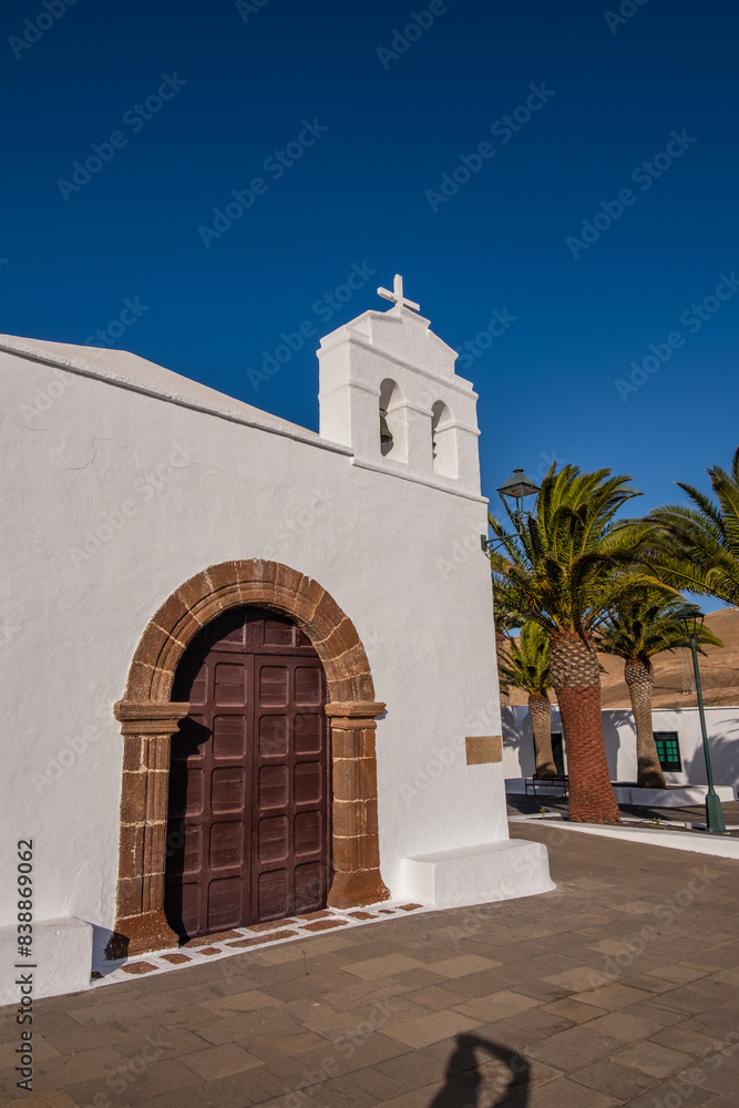 Fototapeta premium view of the main facade of the church of the village of Femés. Brown wooden door. Row of palm trees. Town square. Clear sky. Lanzarote, Canary Islands, Spain.
