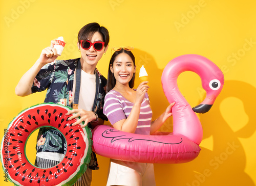 Smiling Asian couple with a mobile phone and a rubber ring, wearing t-shirts featuring coconut tree flowers, vibrant yellow backdrop, summer,