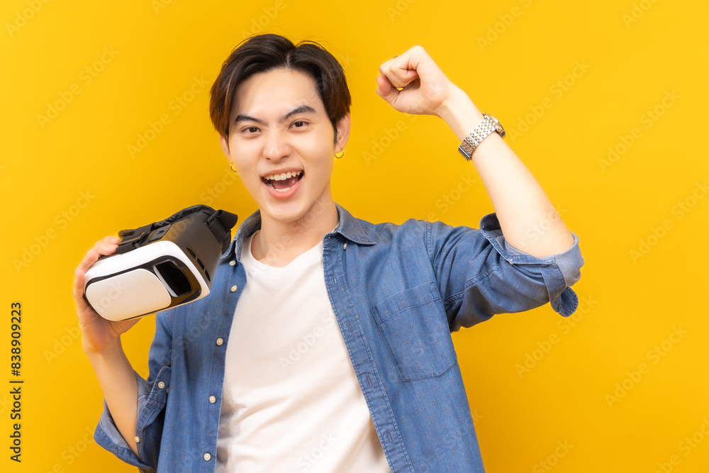 Asian young man in jacket jean and playing video games using vr with headphones on yellow background isolated. Asian young man in jacket jean and playing video games using vr with headphones on yellow background isolated.