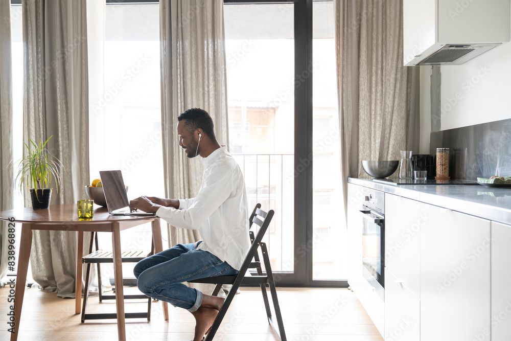 Smiling man sitting at table in modern apartment using laptop and earphones