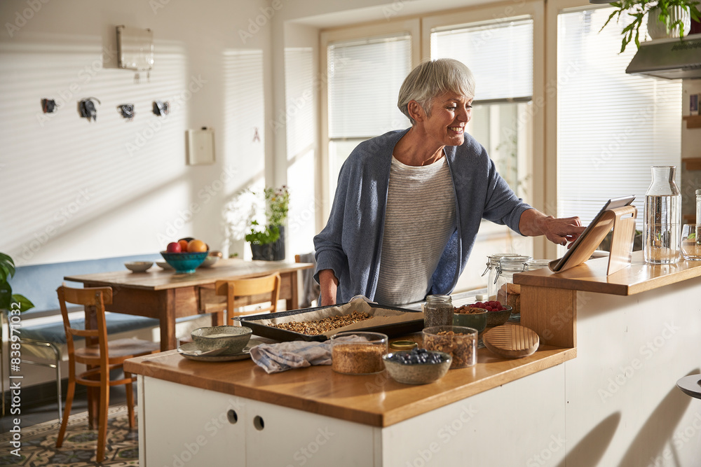 Smiling senior woman using digital tablet in kitchen while preparing granola Smiling senior woman using digital tablet in kitchen while preparing granola