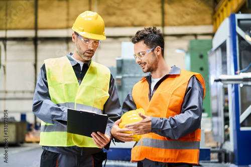 Two men wearing protective workwear holding clipboard and talking in factory