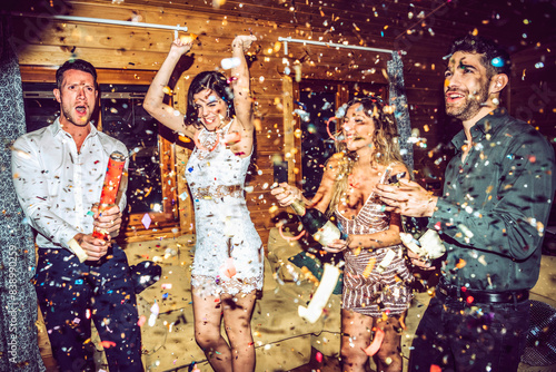 Happy friends opening champagne while dancing amidst confetti in party