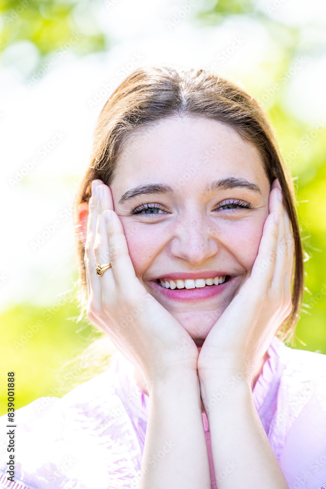 This portrait captures a young woman holding her face in her hands, her eyes crinkling with a broad, beaming smile. The sunlight adds a radiant quality to her complexion, and the out-of-focus natural This portrait captures a young woman holding her face in her hands, her eyes crinkling with a broad, beaming smile. The sunlight adds a radiant quality to her complexion, and the out-of-focus natural