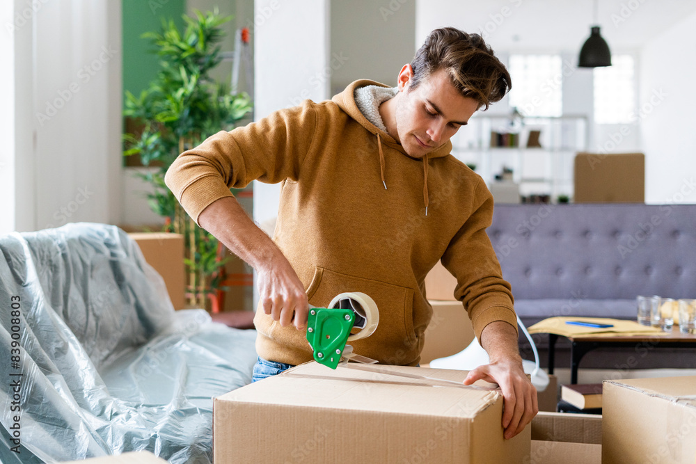 Young man applying adhesive tape on cardboard box while moving in new home