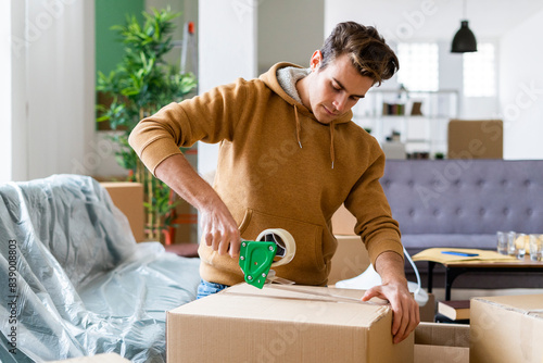 Obraz na płótnie Young man applying adhesive tape on cardboard box while moving in new home w salonie