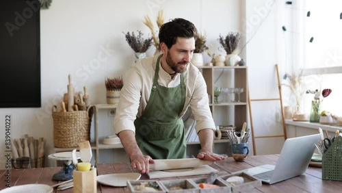 Mature bearded male potter flattens clay with rolling pin to create handmade dishes. Process of making piece of pottery art in bright studio against background of shelves with products. photo