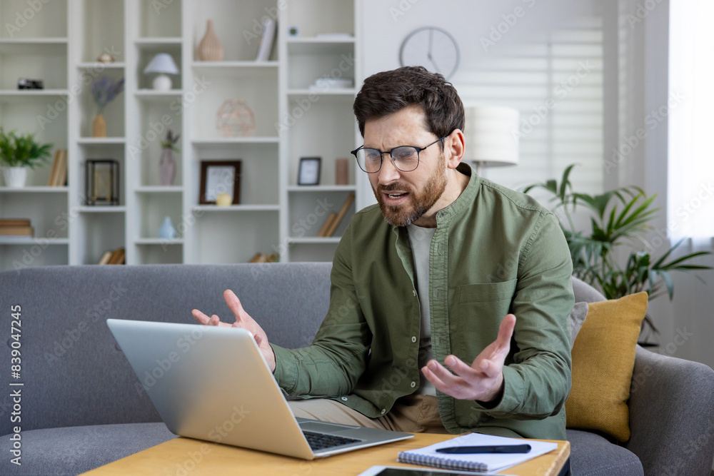 Frustrated man in casual wear gesturing during a video call from his home office, expressing stress and confusion while using a laptop Frustrated man in casual wear gesturing during a video call from his home office, expressing stress and confusion while using a laptop