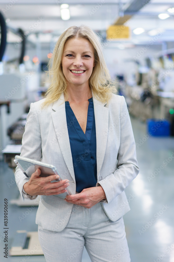 Smiling mature blond businesswoman holding digital tablet while standing at illuminated industry Smiling mature blond businesswoman holding digital tablet while standing at illuminated industry