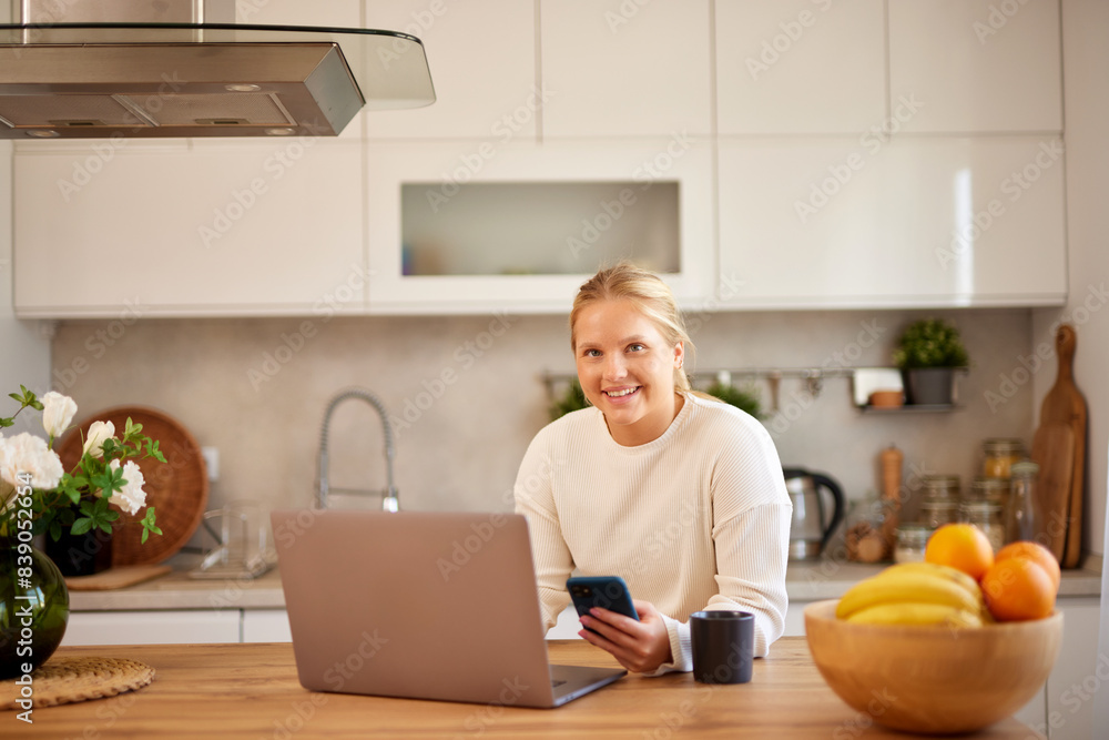 Young woman using mobile phone while working on laptop in the kitchen Young woman using mobile phone while working on laptop in the kitchen