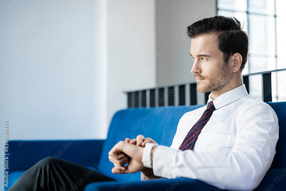 Businessman sitting on blue couch in office checking smartwatch Businessman sitting on blue couch in office checking smartwatch