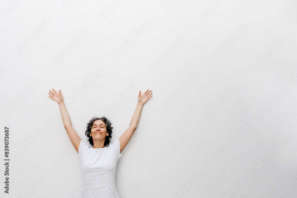 Smiling mature woman standing with eyes closed and arms raised against white wall Smiling mature woman standing with eyes closed and arms raised against white wall