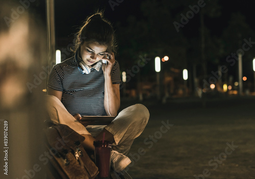 Young woman with smoothie sitting on bench at night using tablet and headphones photo