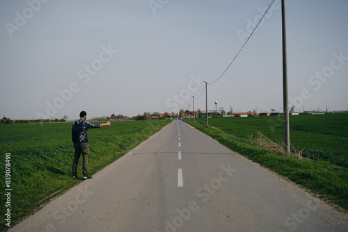 Travel concept. Young man hitchhiker with backpack  stands by rural country road on sunny spring day and waiting for car. Front view. Guy went on trip alone  trying to stop passing car. Rear view.