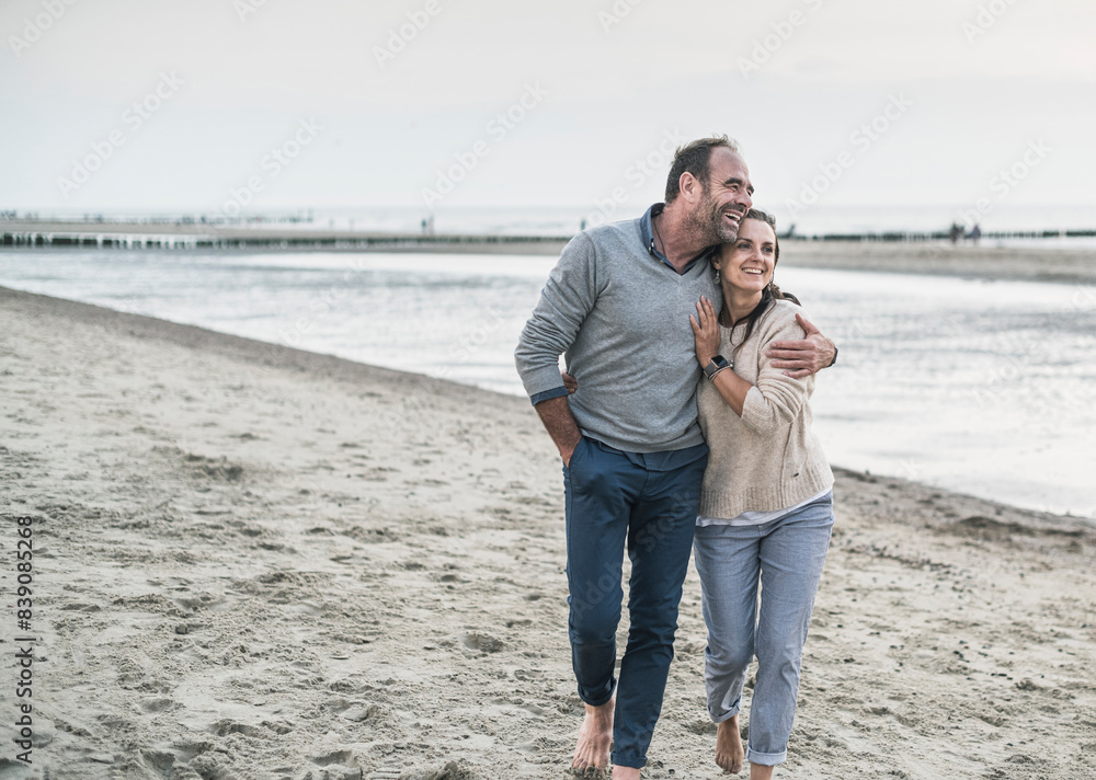 Happy mature couple embracing while walking at beach against sky during sunset