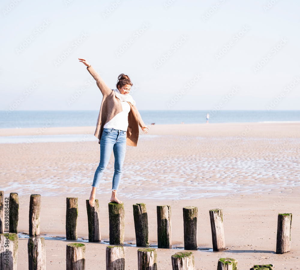 Smiling young woman with arms outstretched walking on wooden posts at beach during sunny day