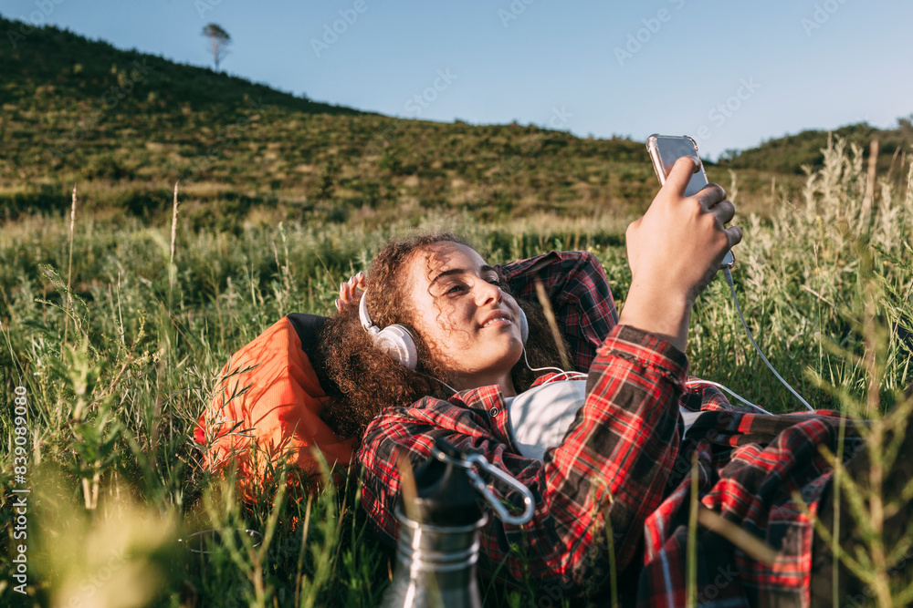 Teenage girl with backpack lying on a meadow listening music with headphones and cell phone