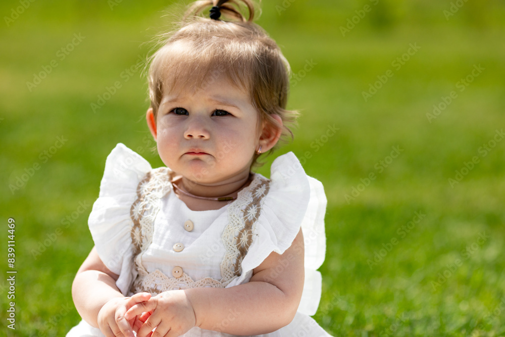A baby in a white dress sitting on the grass and crying. crying baby toddler girl on grass outdoors. Child feelings and emotions concept. Sad expression. Childhood. copy space