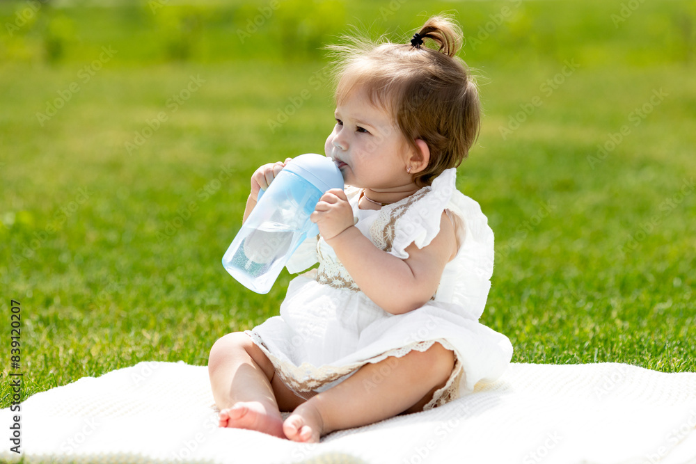 A cute toddler girl in a white summer dress sitting on the grass in the summer, drinking water from a bottle. Healthy toddler one year sitting on the grass drinking water from a bottle A cute toddler girl in a white summer dress sitting on the grass in the summer, drinking water from a bottle. Healthy toddler one year sitting on the grass drinking water from a bottle