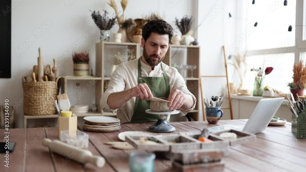 Smiling man working with clay in cozy home pottery studio. Bearded male seated at wooden table with various tools, materials, and laptop in bright well-decorated room. Smiling man working with clay in cozy home pottery studio. Bearded male seated at wooden table with various tools, materials, and laptop in bright well-decorated room.