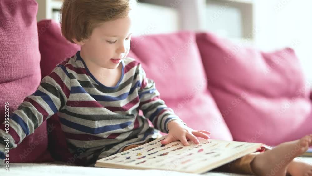 Cute toddler boy reading books on the sofa at home on a bright sunny afternoon. Daytime care creative activity. Kids having fun. Educational learning games. Family leisure indoor. Cute toddler boy reading books on the sofa at home on a bright sunny afternoon. Daytime care creative activity. Kids having fun. Educational learning games. Family leisure indoor.