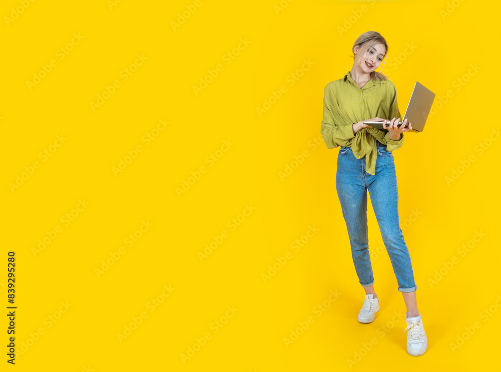 Using laptop, full body caucasian young 20s blonde student or employee or businesswoman using laptop. Focused cheerful girl browsing internet, typing on yellow background. Copy space. Using laptop, full body caucasian young 20s blonde student or employee or businesswoman using laptop. Focused cheerful girl browsing internet, typing on yellow background. Copy space.
