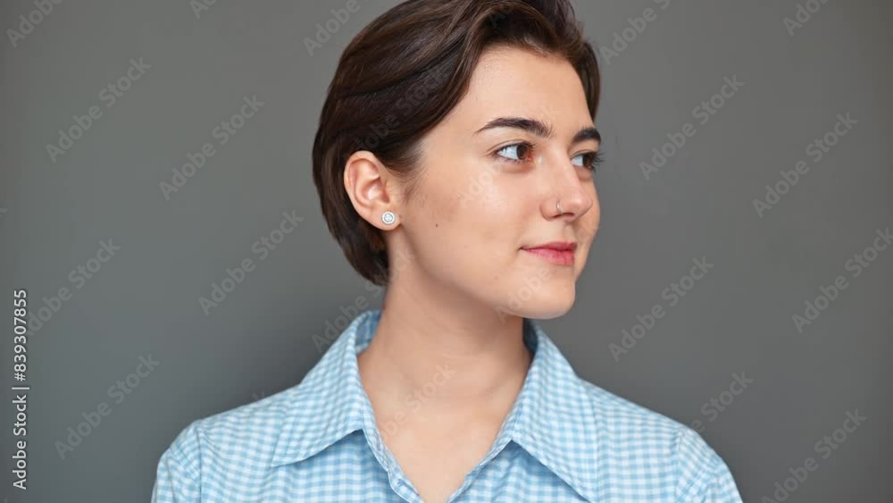 Portrait of smiling cute young brunette woman with short haircut in blue shirt turning around and looking at camera standing on grey background Portrait of smiling cute young brunette woman with short haircut in blue shirt turning around and looking at camera standing on grey background